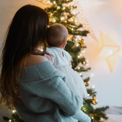 Back view of anonymous mother standing with little baby on hands and looking at Christmas tree with festive decorations and garlands at home